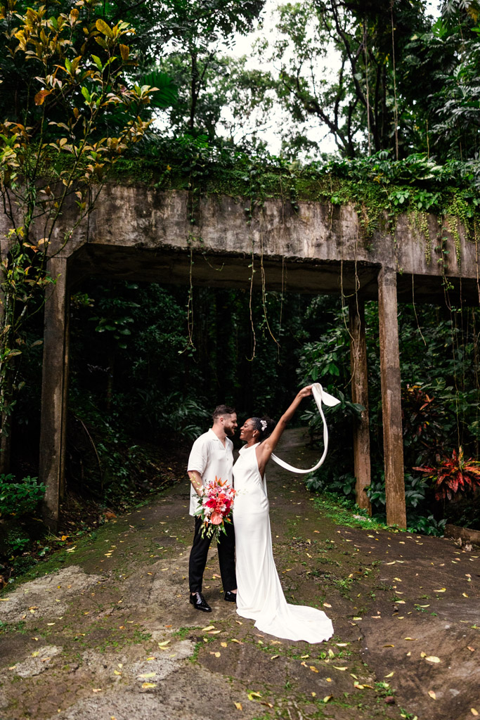 A bride and groom stand under a concrete waterway in the lush forest at Diamond Falls Botanical Gardens