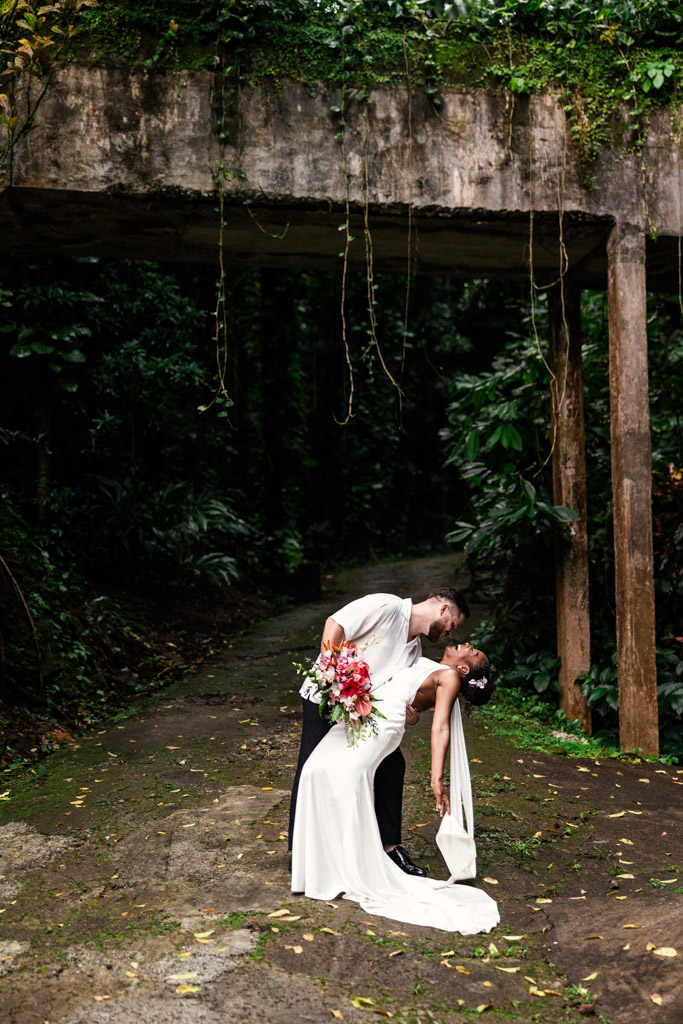 A groom dips and kisses his bride on a mossy path under a rustic concrete bridge surrounded by lush greenery at Diamond Falls Botanical Gardens