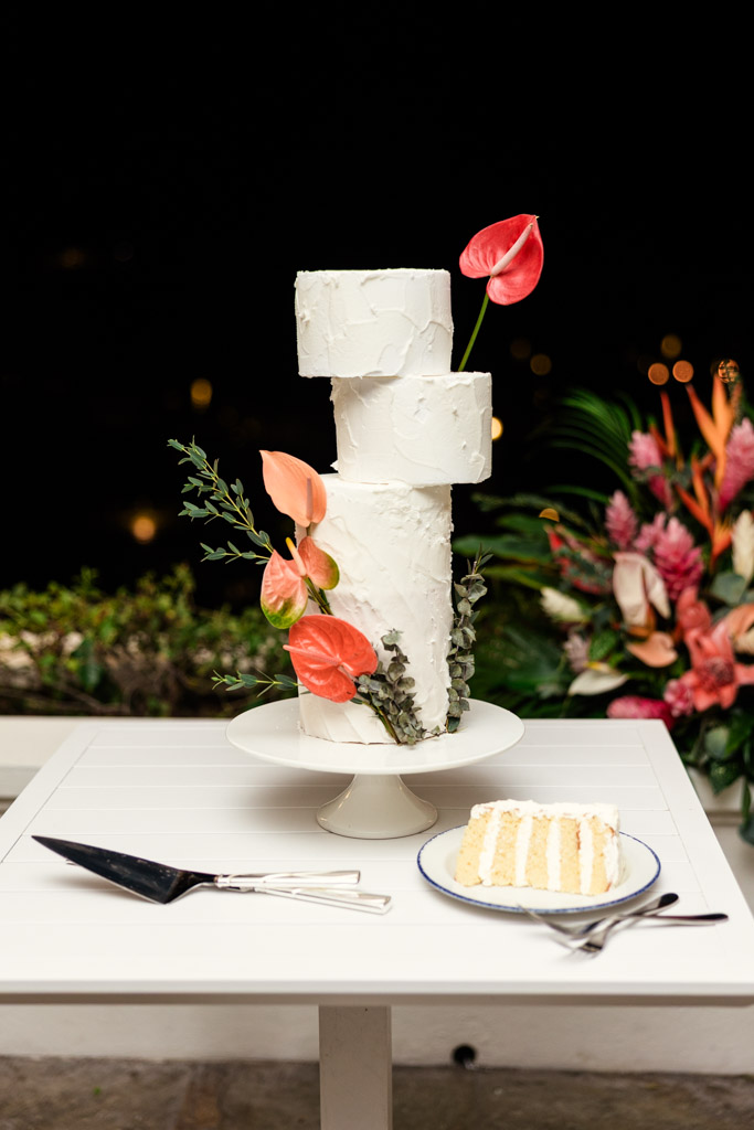 Three-tier white cake with pink anthurium flowers on a stand with a slice on a plate in front for Viceroy Sugar Beach wedding celebration