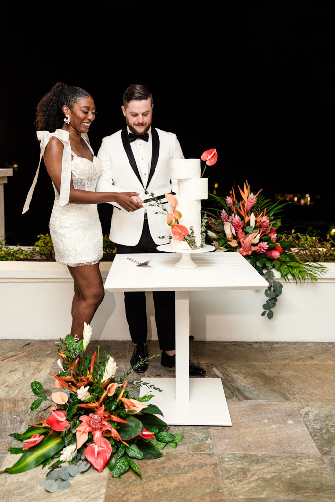 Newlywed couple in formal attire cuts a white wedding cake decorated with tropical flowers at Viceroy Sugar Beach