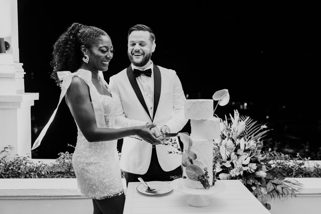 Black and white photo of smiling newlyweds in formal attire cuts a tall wedding cake together at their Viceroy Sugar Beach outdoor nighttime celebration