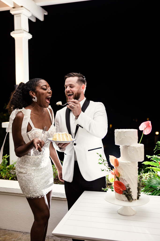 Newlyweds in formal attire laugh while feeding each other wedding cake at night, celebrating their special day at Viceroy Sugar Beach