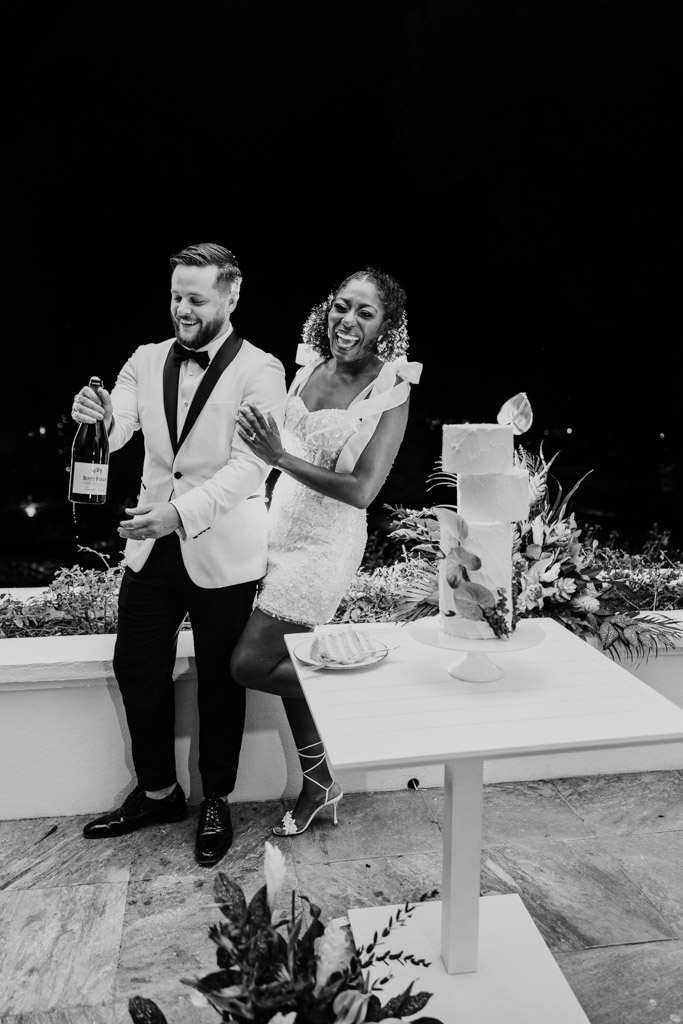 Black and white photo of joyful couple celebrating with champagne beside a decorated cake at their wedding reception at Viceroy Sugar Beach