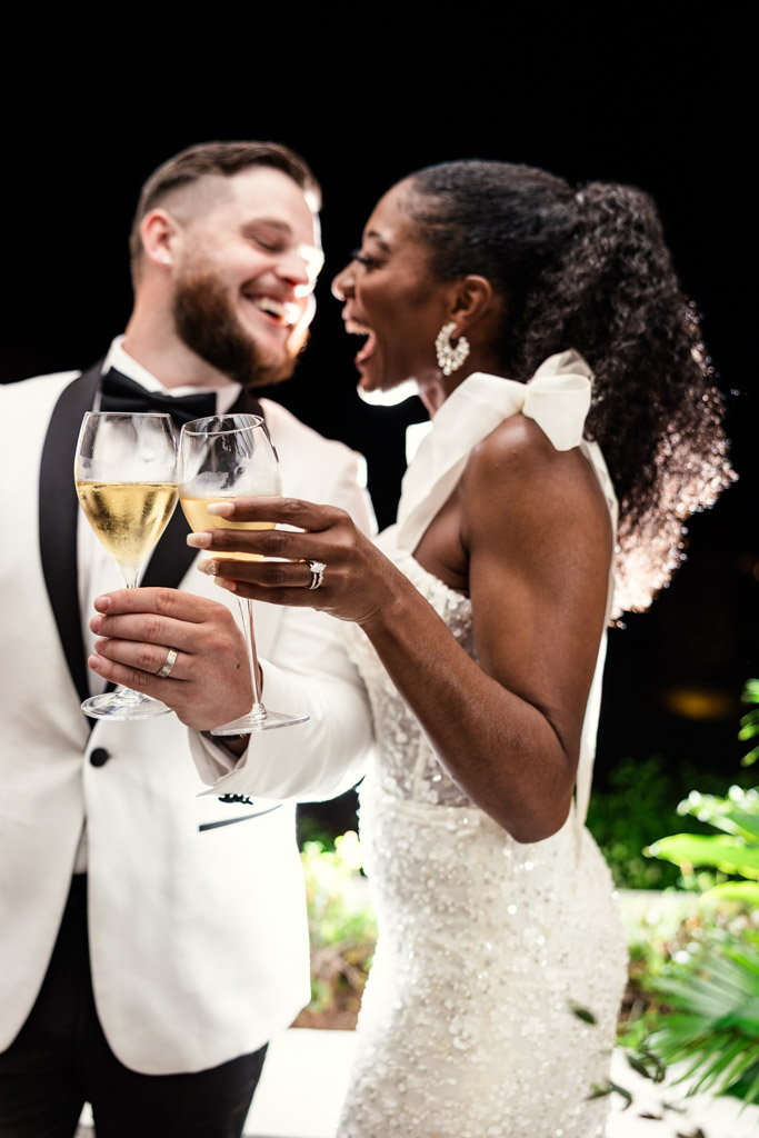 Happy bride and groom in formal attire laugh and toast with champagne at night at Viceroy Sugar Beach