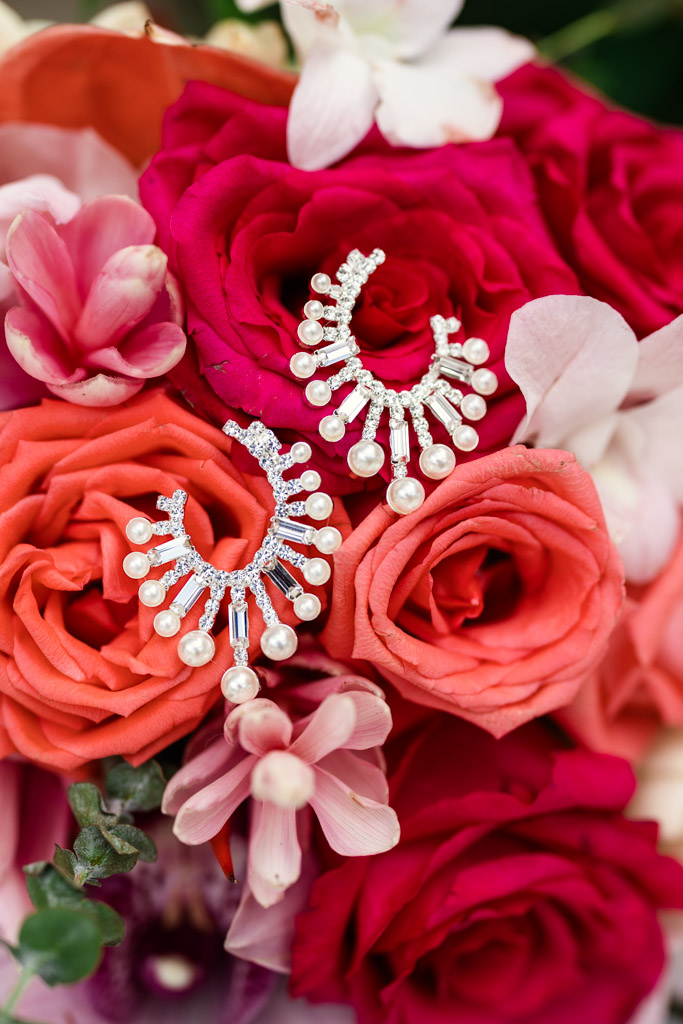 Detail photo of diamond and pearl earrings placed on a bouquet of vibrant pink, red, and orange roses with orchids for Viceroy Sugar Beach wedding celebration
