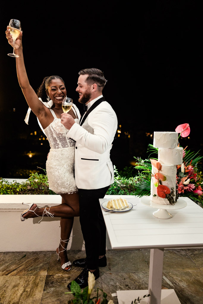 Bride and groom in formal attire celebrate with drinks next to a white wedding cake at night, during Viceroy Sugar Beach wedding celebration