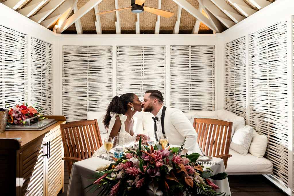 Newlywed couple kisses at a table set for two, surrounded by flowers in a cozy, white cabana at Viceroy Sugar Beach