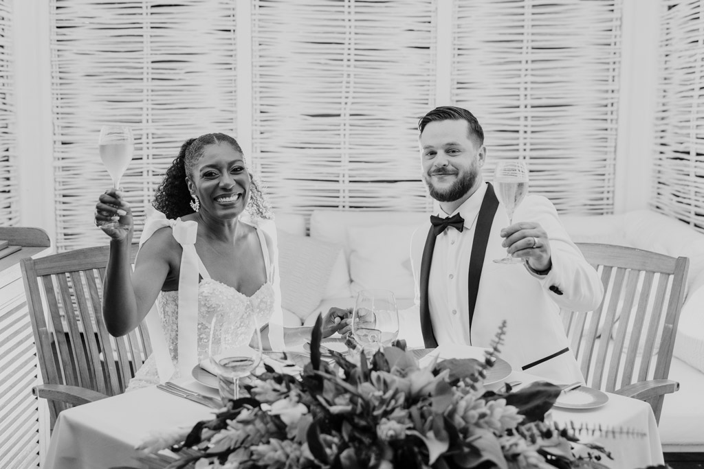 Black and white photo of newlyweds in formal attire raising glasses at a decorated table, celebrating together at Viceroy Sugar Beach