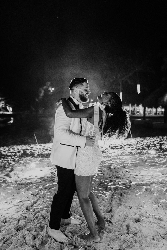 Black and white photo of barefoot newlywed couple in formal attire dancing and smiling on the sandy shores of Viceroy Sugar Beach at night