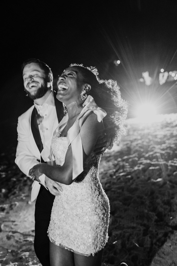 Black and white photo of happy newlywed couple in formal attire laughing together on the moonlit sands of Viceroy Sugar Beach