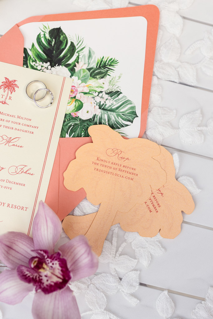 Wedding invitation suite with floral designs, rings, and an RSVP card shaped like a palm tree, next to a pink flower for a Viceroy Sugar Beach wedding celebration