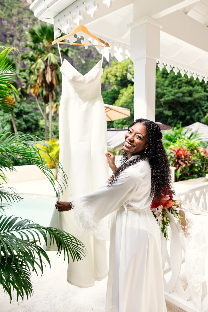 Smiling bride in a robe holds a white wedding dress on a hanger at Viceroy Sugar Beach