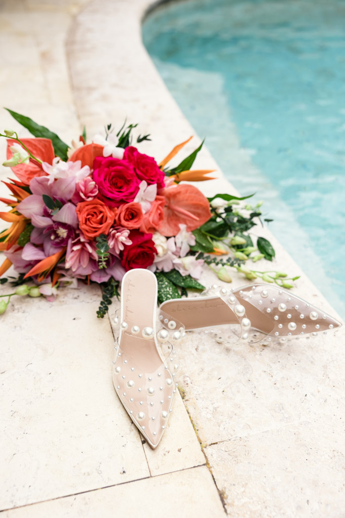 Bride's pearl-studded heels and a vibrant bouquet of flowers placed by the edge of a swimming pool at Viceroy Sugar Beach