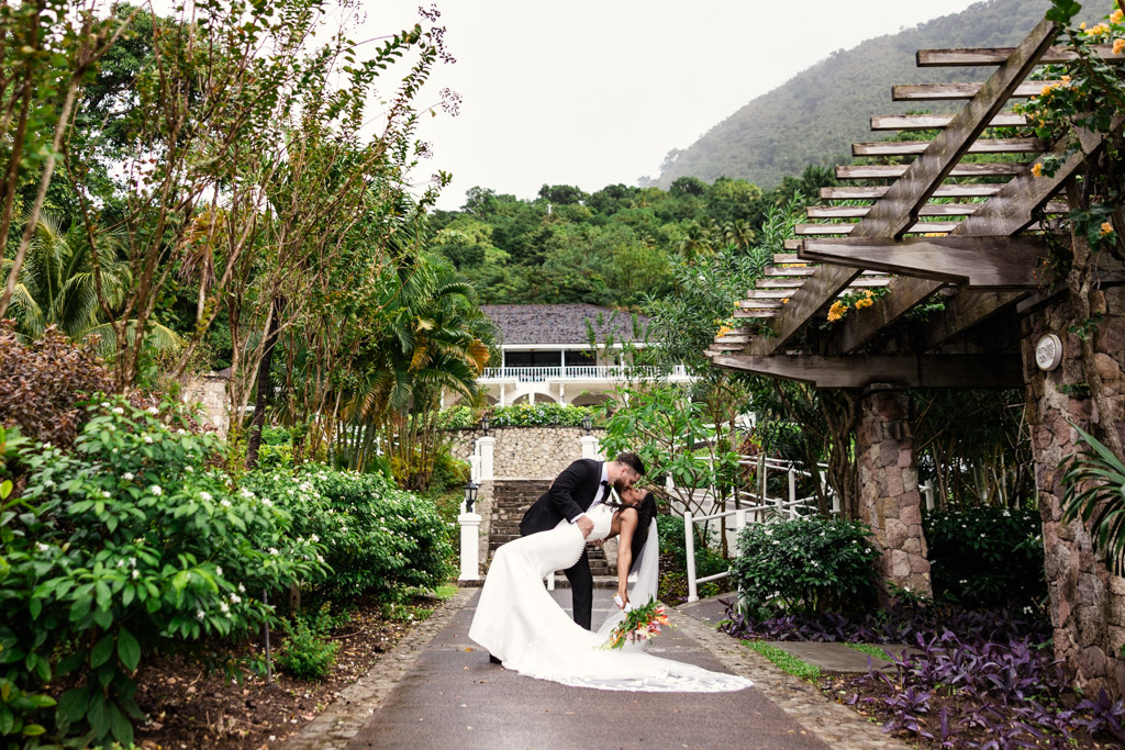 A groom dips and kisses his bride on a garden path with lush greenery and a wooden pergola at Viceroy Sugar Beach