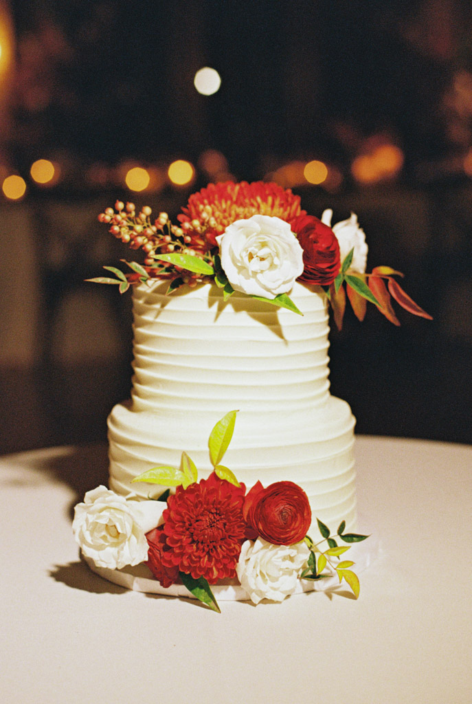 Film photo of two-tier white wedding cake adorned with red and white flowers, set on a table with a dark background for Cafe Brauer wedding celebration