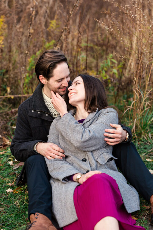 A couple sits on grass, smiling and embracing each other in an outdoor, autumn setting.
