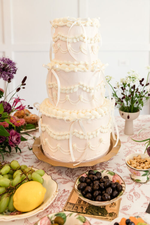 Three-tiered white cake with ornate icing and ribbons, surrounded by fruit, nuts, and flowers on a table.