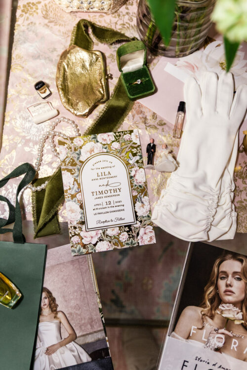 Floral wedding invitation surrounded by gloves, jewelry, perfume, magazine, and green velvet ribbon on a table.