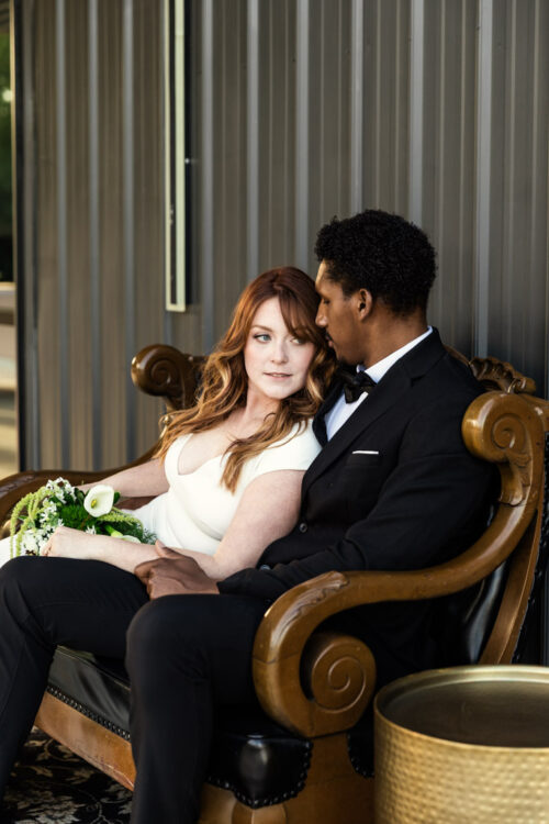 A couple dressed formally sits close together on a vintage sofa, holding hands and gazing at each other.