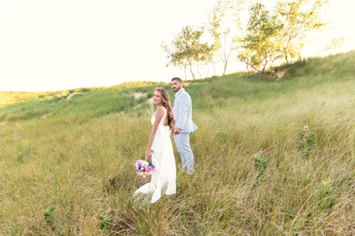 Bride and groom stand together in a grassy field, the bride holding a bouquet and looking back.