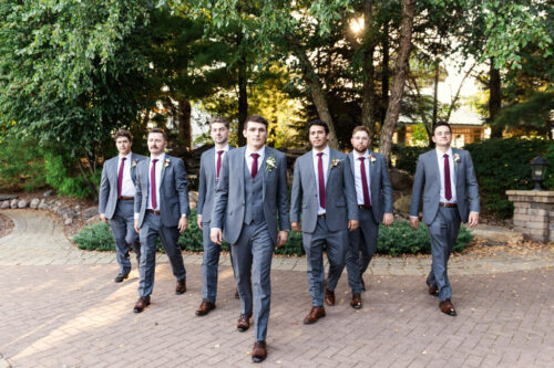 Seven men in grey suits with red ties walk confidently outdoors on a sunny, tree-lined path.