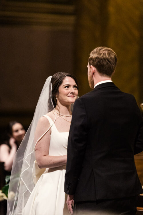 Bride and groom face each other, smiling, during their wedding ceremony indoors.