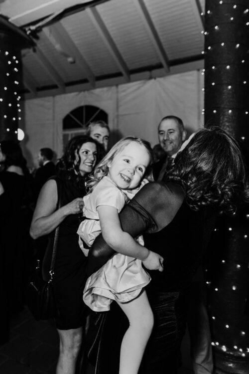 A smiling child in a dress is held by a woman at a lively indoor event with people in the background.