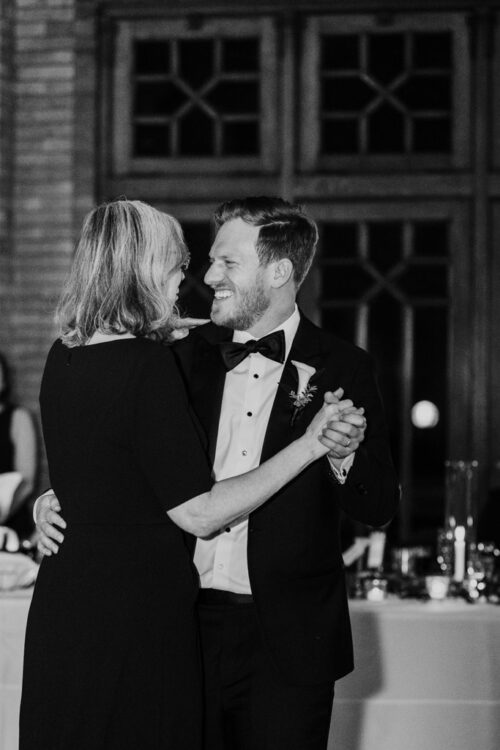 A smiling man in a tuxedo dances with an older woman at a formal event.