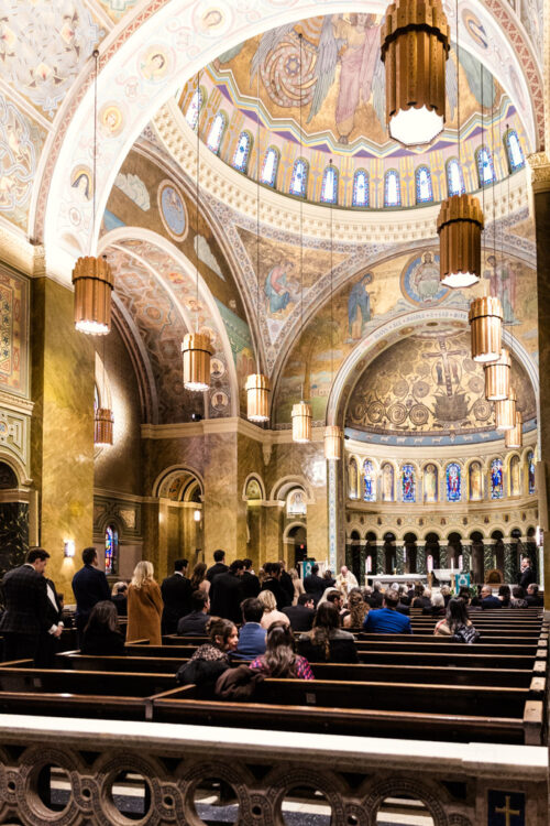 People seated in pews inside an ornate church with arched ceilings and colorful murals.