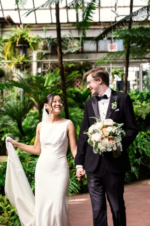 A bride and groom smile at each other, holding hands, surrounded by lush greenery in a greenhouse.