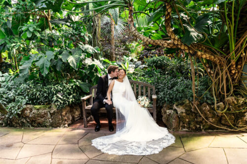 Bride and groom sit on a bench in a lush garden, surrounded by greenery and tropical plants.