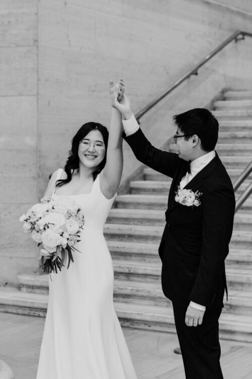 A bride and groom smile as he lifts her arm; she holds a bouquet and wears a flowing white dress.