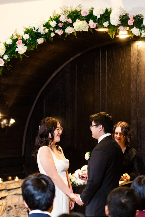 A bride and groom hold hands and smile at each other during their indoor wedding ceremony under floral decorations.