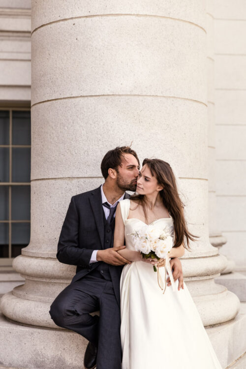 A groom kisses his bride's head as they sit together outside, with the bride holding a bouquet of white roses.