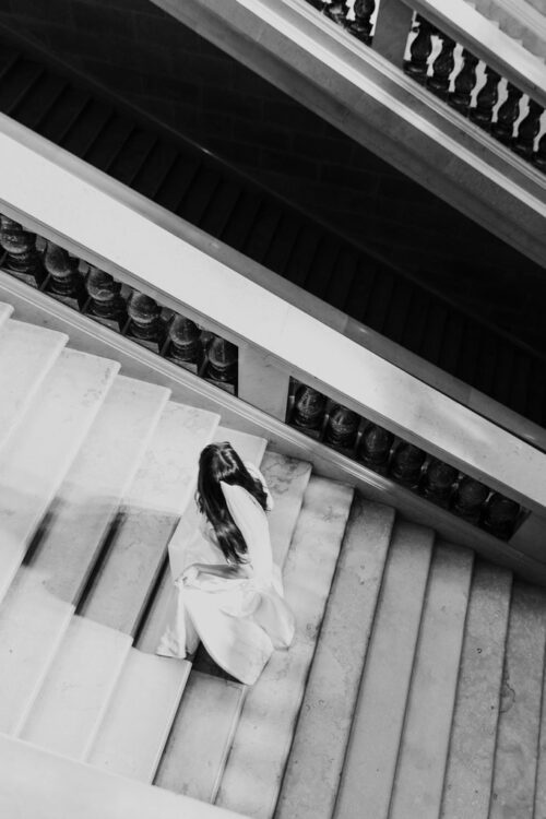A woman in a long dress sits alone on a staircase in a dramatic, black-and-white setting.