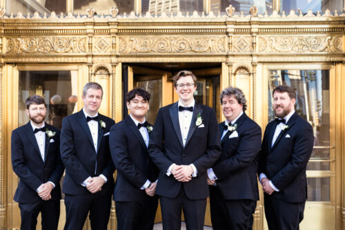 Six men in black tuxedos stand smiling in front of ornate gold doors at a formal event.