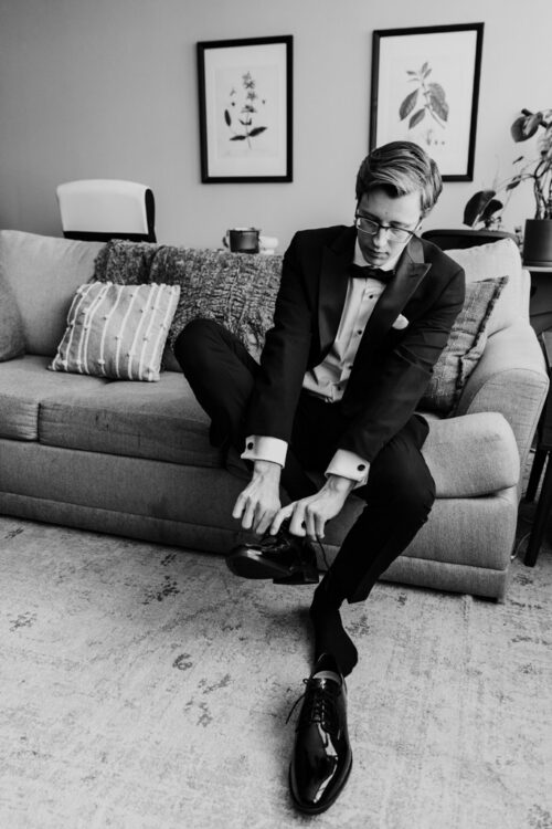 A man in a tuxedo sits on a couch, putting on his shoe, with framed artwork on the wall behind him.