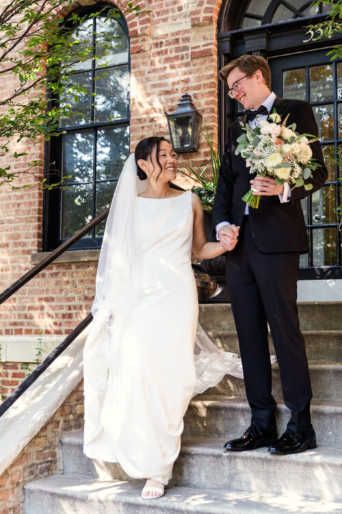 A bride and groom smile at each other while holding hands on outdoor steps in wedding attire.