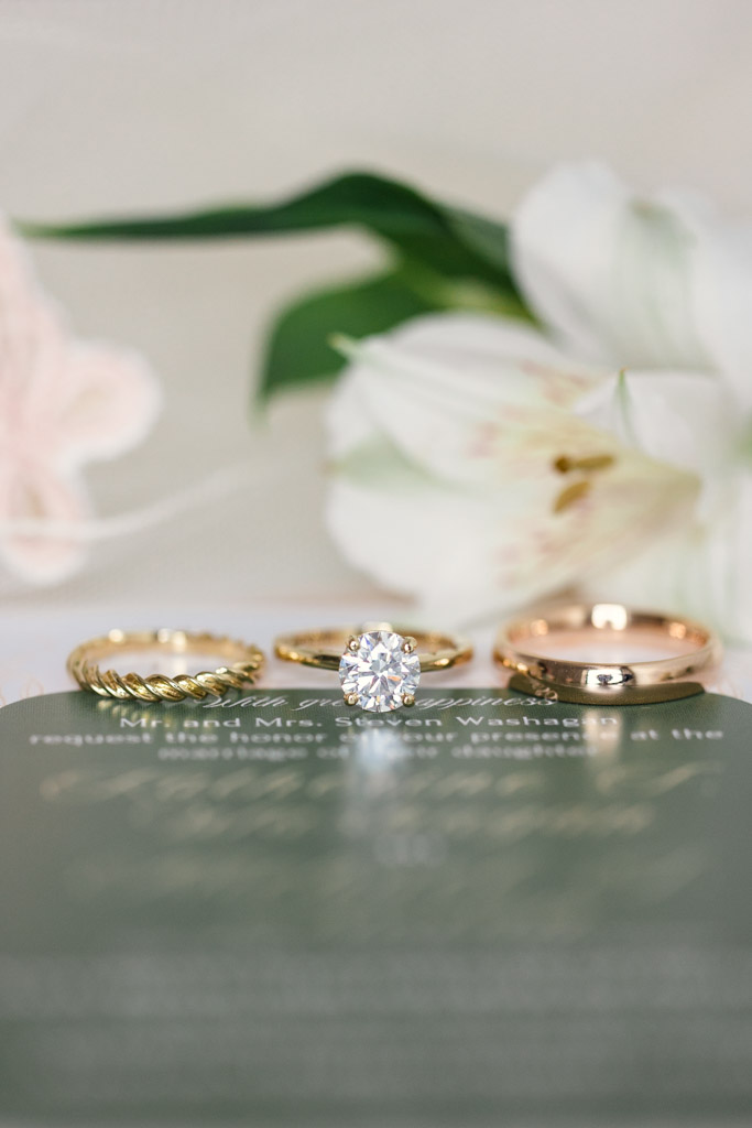 Three gold rings, including a diamond engagement ring, displayed in front of white flowers on a green wedding invitation