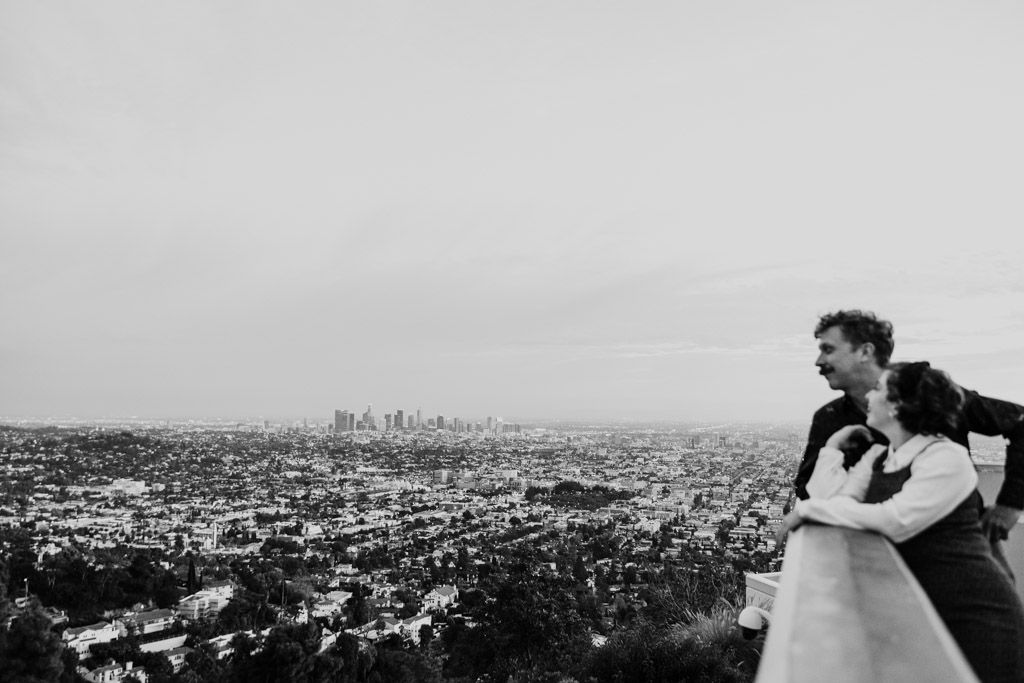 Black and white photo of couple standing on a balcony at Griffith Park Observatory overlooking Los Angeles with skyscrapers in the distance, under a cloudy sky
