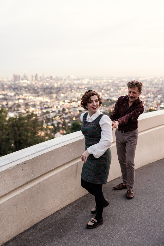 A smiling couple walks playfully along a wall at Griffith Park Observatory with Los Angeles cityscape blurred in the background