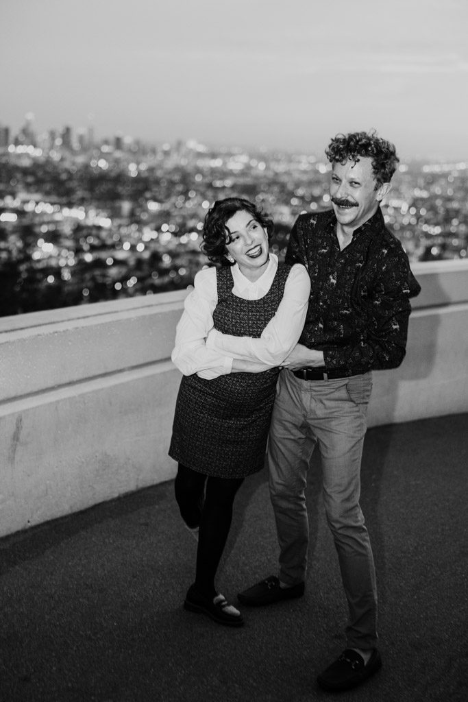 Black and white photo of happy engaged couple posing together at Griffith Park Observatory with Los Angeles cityscape blurred in the background