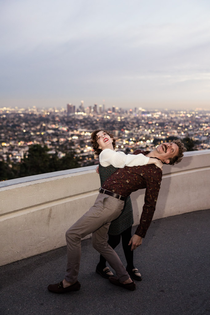 Two people pose playfully at Griffith Park Observatory at dusk, laughing and leaning dramatically