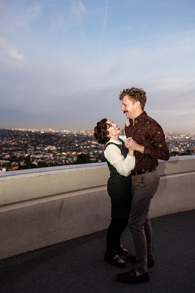 Engaged couple dances and laughs together near Griffith Park Observatory at sunset with Los Angeles in the background