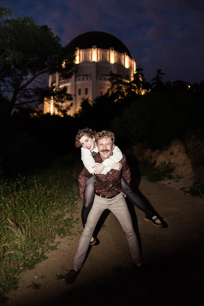 A man gives a woman a piggyback ride at night near the illuminated Griffith Observatory