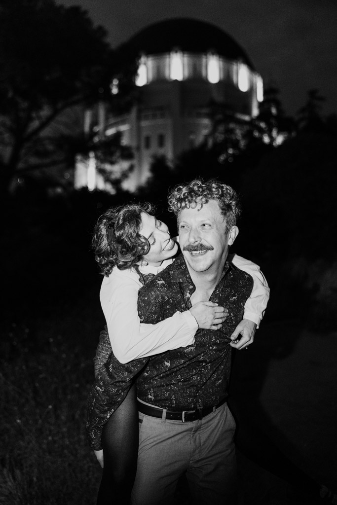 Black and white photo of woman riding on her partner's back, both smiling, with Griffith Park Observatory illuminated behind them at night