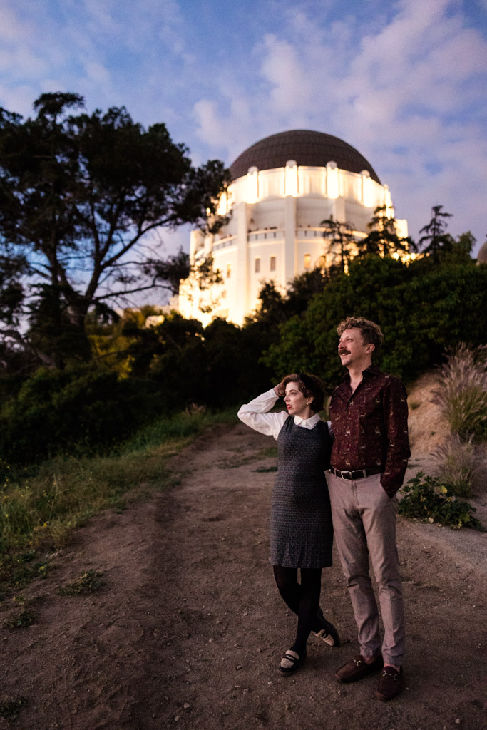 Engaged couple stands on a dirt path near Griffith Park Observatory at dusk, surrounded by trees and greenery