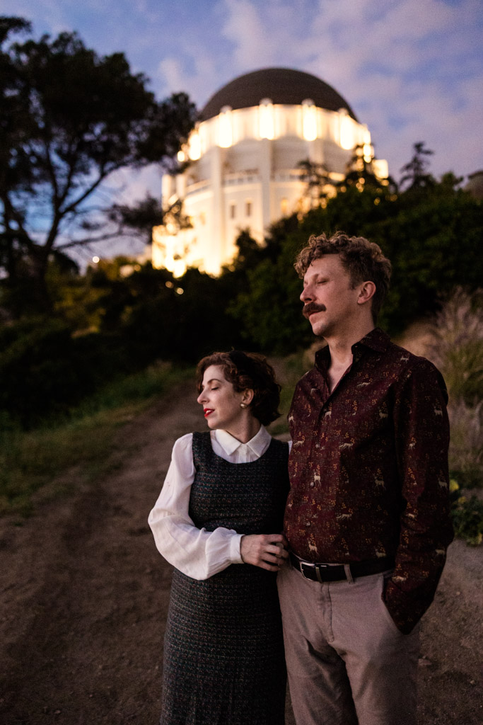 A couple stands on a path at dusk with the Griffith Park Observatory lit up in the background