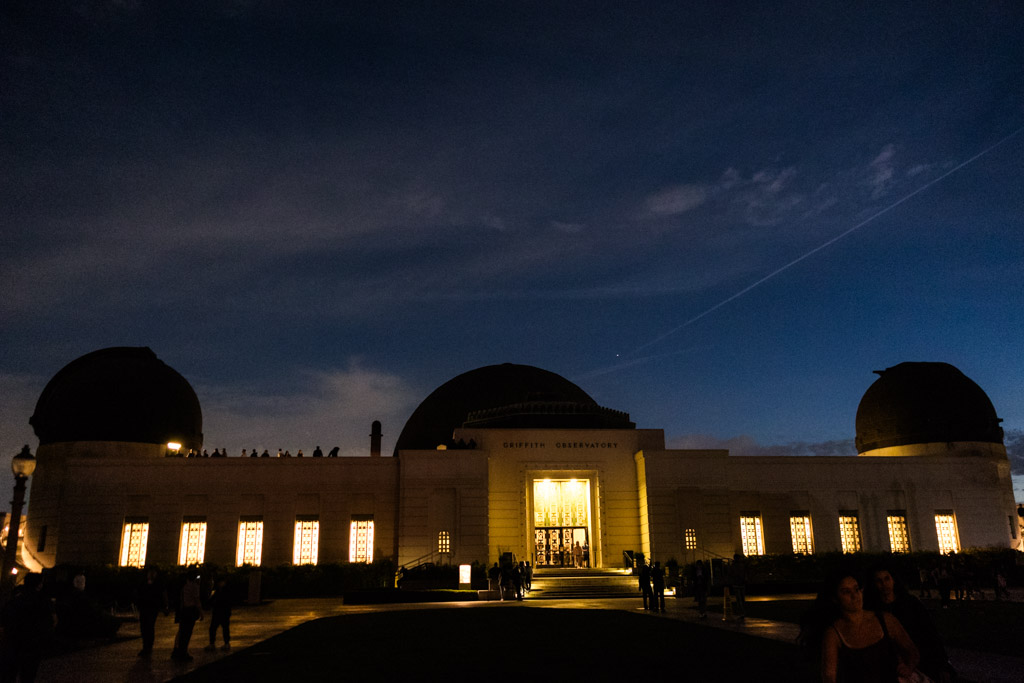 Griffith Park Observatory at night, warmly lit, with silhouettes of people outside under a dark blue sky