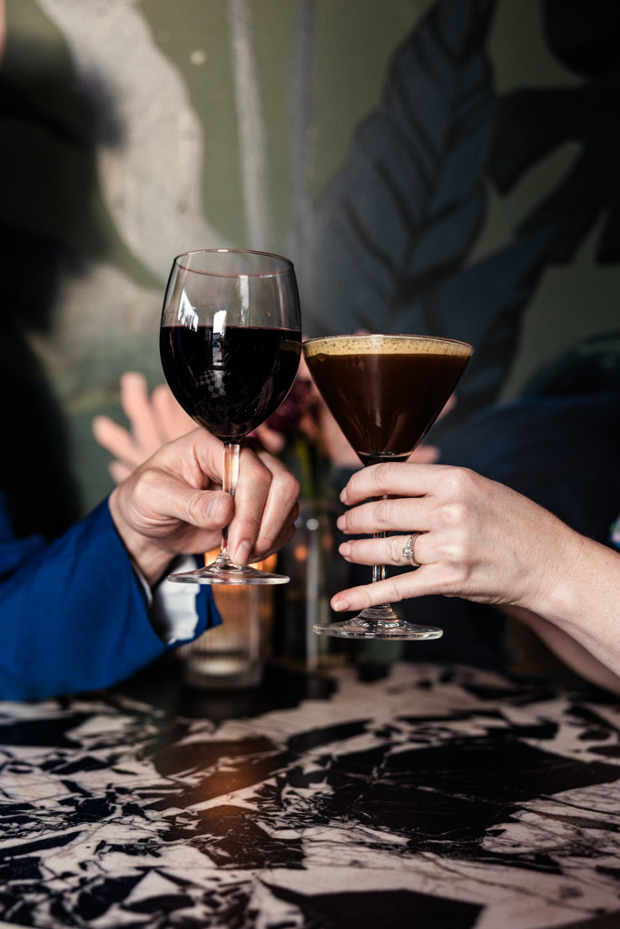 Engaged couple toasts a glass of red wine and a cocktail at a marble table in Justine's